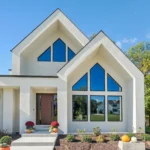 Modern white house with large triangular windows, a wooden front door, and a neatly landscaped yard decorated with pumpkins and potted flowers. Trees and blue sky are visible in the background.