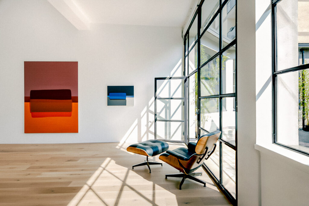 A bright, modern room with Inicio window + doors, wooden floors, an Eames lounge chair and ottoman, and two abstract paintings on a white wall. Sunlight streams in, casting soft shadows across the floor and furniture.