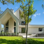 A modern white house with geometric architecture, large overhangs, expansive windows, and a patio area, surrounded by green grass, trees, and blue sky.
