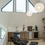 A modern kitchenette features wooden cabinets, a sink, coffee machines, and a lounge chair with a matching ottoman in black and white patterns, set beneath large triangular windows and round pendant lights.