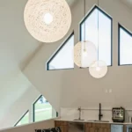 Modern kitchen interior with triangular windows, three hanging spherical pendant lights, a sink, coffee machine, and light wood cabinetry under a high sloped ceiling. Natural light brightens the room.