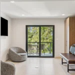 Modern, minimalist living space with a gray swivel chair, a wooden bench with gray cushions, large tiled floor, and glass doors opening to a balcony with outdoor seating and views of green trees.
