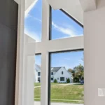 Large, modern windows with geometric shapes let in natural light and offer a view of green grass, blue sky, and white houses outside. A few small pumpkins sit on the windowsill inside.