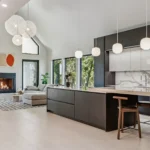 Modern open-concept kitchen and living room with large windows, dark cabinets, marble backsplash, pendant lights, a sectional sofa, fireplace, and contemporary decor in a bright, airy space.