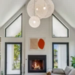 A modern living room with vaulted ceilings, large triangle windows, a lit fireplace, a gray sectional sofa, geometric artwork, globe pendant lights, a striped rug, and greenery outside visible through glass doors.