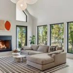 A modern living room with large black-framed windows, a beige sectional sofa on a checkered rug, a round coffee table, a lit fireplace, abstract wall art, and views of trees outside.