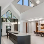 Modern open-concept kitchen and dining area with high vaulted ceilings, large geometric windows, minimalist dark cabinetry, a central island, pendant lights, and a long dining table with chairs.