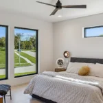 A modern bedroom with large windows, a gray bed with striped bedding, a yellow pillow, a round nightstand, and a ceiling fan. Sunlight fills the room, and green lawns and trees are visible outside.
