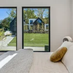 A bright bedroom with a neatly made bed, gray headboard, and two large windows showing a view of modern black houses, green grass, and trees outside on a sunny day.