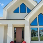 Modern house with white exterior, large geometric windows, and a wooden front door. Steps to the entrance are decorated with red potted flowers and various pumpkins. The sky is clear and blue.