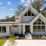 A modern white house with large triangular windows, sloped rooflines, and neatly landscaped front yard. A walkway with pumpkins and potted plants leads to the entrance. Trees and a clear sky are in the background.