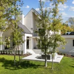 Modern white two-story house with large windows, angular rooflines, and a covered patio, surrounded by green grass, trees, and a landscaped yard under a blue sky with scattered clouds.