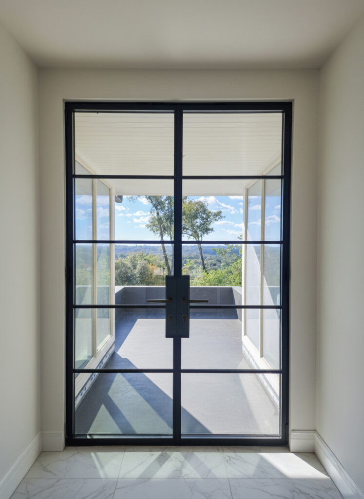 A large INICIO steel window + doors with a black metal frame opens to a balcony with a scenic view of trees, hills, and blue sky, seen from a bright, modern interior with white walls and marble flooring.