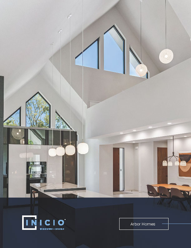 Modern home interior with high ceilings, large angular windows, and pendant lighting over a kitchen island. The space features white walls, dark accents, and a dining area with contemporary chairs—reflecting the style found at the INICIO showroom.