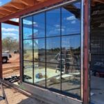 Large window with black grid panes installed in a partially constructed building; construction materials and ladders are visible inside and outside, with a reflection of the blue sky and landscape in the glass.