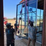 Three workers install a large glass window panel with suction cup equipment inside a wooden building frame, while using red straps to lift the glass. The outdoor scene is visible through the opening.