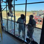 Three people are installing a large glass window; one stands on a ladder outside, while two others stand on the ground holding the glass with suction tools. Construction equipment and a field are visible in the background.