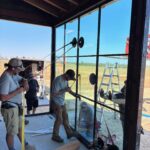 Four workers install a large glass window pane using suction cups and support tools inside a partially built structure. A ladder and trailer are visible outside on a sunny day.
