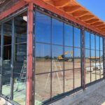 Large glass windows with black metal frames are installed on the corner of a building under construction. Construction equipment and a ladder are visible outside, and blue sky can be seen in the background.