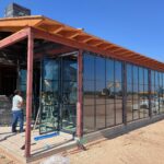 A building under construction with large glass windows being installed. A worker stands near the glass, tools are scattered on the ground, and a yellow excavator is visible in the background under a clear blue sky.