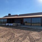 A partially constructed building with large glass windows, a covered porch, and roofing in progress. Construction materials and tire tracks are visible in the dirt in front of the building under a clear blue sky.