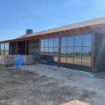 A building under construction with large floor-to-ceiling glass windows, unfinished walls, a pile of stone blocks nearby, and a person wearing a hat and mask walking in front. The sky is clear and sunny.