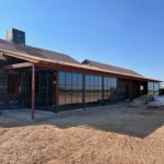 A single-story house under construction with large glass windows and doors, an unfinished roof, and exposed black house wrap. Construction materials are scattered around the dirt ground. The sky is clear and blue.