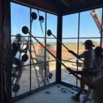 Several workers use suction cup tools to install a large glass window pane in a building under construction. The window offers a view of a sunny, open landscape with dirt and construction equipment outside.